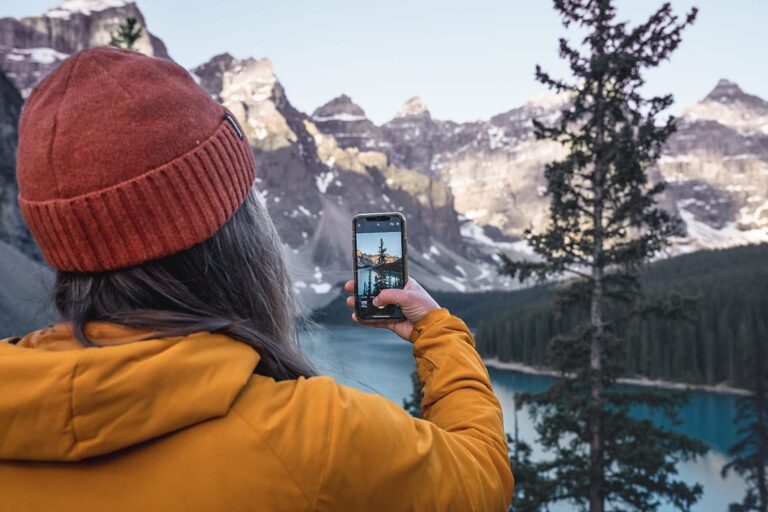 The back of a personâ€™s head, theyâ€™re holding up a phone and taking a photo of a tree by a lake
