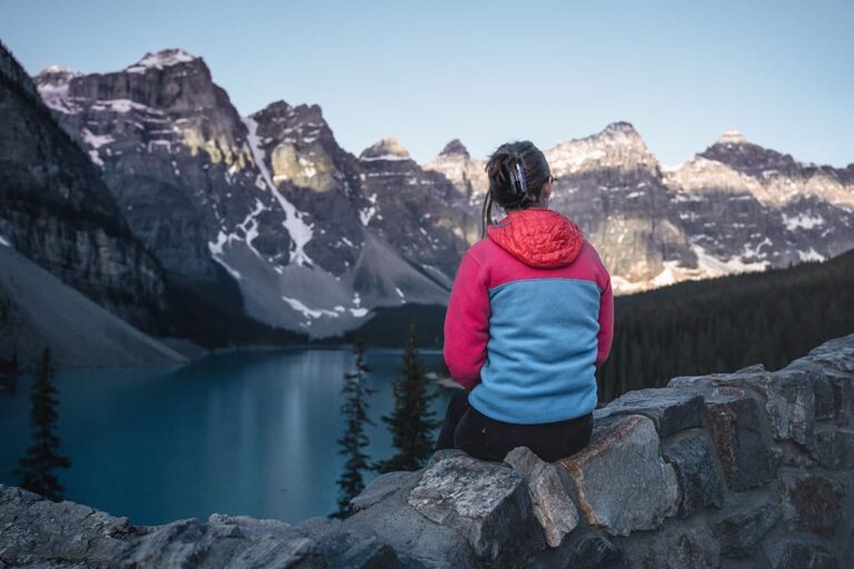 The back of a woman in a pink and blue fleece; she's sitting on a rock wall looking at a lake